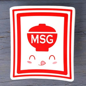 May include: A white sticker with a red border and a red bowl graphic with the letters "MSG" inside. The bowl has a smiling face with a tongue sticking out and pink cheeks.