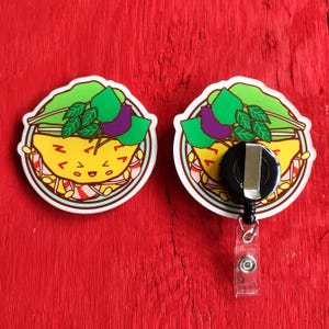 May include: Two circular badge reels featuring a colorful illustration of a smiling yellow omelet topped with green lettuce and purple leaves. One badge reel shows the front graphic, while the other displays the black plastic retractable mechanism.