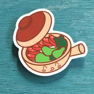May include: A die-cut sticker featuring a cartoon clay pot with a lid tilted open. The pot is light tan with a smiling face, containing red sliced vegetables and green leafy greens. The design has a thick white border and a flat, graphic style.