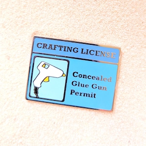 May include: A rectangular pin with a blue background and the words "CRAFTING LICENSE" at the top. The pin features a white glue gun graphic and the text "Concealed Glue Gun Permit."