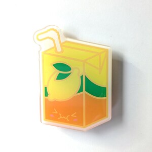 May include: A yellow and orange cartoon juice box with a straw, a lemon, and a green leaf. The juice box has a cute face with a frown and pink cheeks.