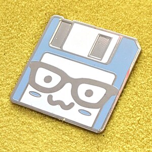 May include: A blue and white enamel pin shaped like a floppy disk with a cartoon face wearing glasses. The face has two large eyes, a small mouth, and two small blush marks on the cheeks.