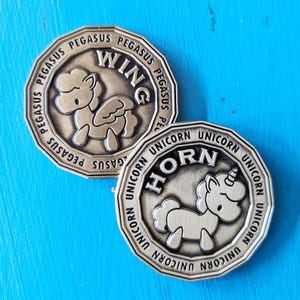 May include: Two gold-colored metal tokens with a unicorn and a pegasus design. The unicorn token has the word "HORN" written on it and the pegasus token has the word "WING" written on it. Both tokens have the word "UNICORN" and "PEGASUS" written around the edge.