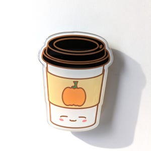 May include: A white coffee cup with a black lid and a pumpkin design on the side. The cup has a cute cartoon face with pink cheeks.