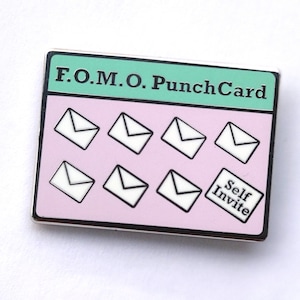May include: A pink and green enamel pin with the text "F.O.M.O. Punch Card" and a graphic of seven white envelopes with black outlines. The bottom right envelope has a small white rectangle with the text "Self Invite" in black.