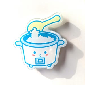 May include: A white and blue cartoon rice cooker with a yellow spoon. The rice cooker has a smiling face with pink cheeks and a square button. The image is a sticker or pin.