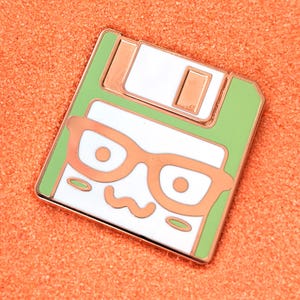 May include: A green and white enamel pin shaped like a floppy disk with a cartoon face wearing glasses. The floppy disk has a copper colored metal slider.