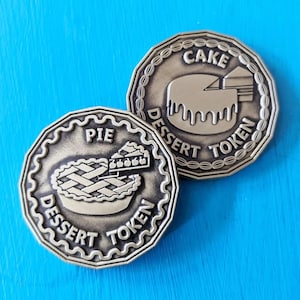 May include: Two bronze-colored dessert tokens with scalloped edges. One features a pie graphic with the text "PIE DESSERT TOKEN". The other shows a cake graphic with the text "CAKE DESSERT TOKEN". Both tokens are on a bright blue background.