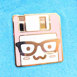May include: A pink and gold enamel pin in the shape of a floppy disk. The pin features a cartoon face with glasses and a smiling mouth. The pin has a gold border and is set against a blue background.