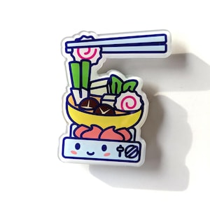 May include: A colorful enamel pin featuring a cartoon illustration of a hot pot with chopsticks, vegetables, and fish cakes. The hot pot is sitting on a small stove with a smiling face and the Japanese character for "fire".