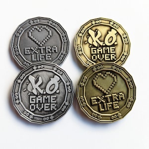 May include: Four silver and gold-colored tokens with pixelated hearts and the text "EXTRA LIFE" and "KO GAME OVER".