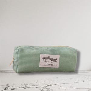 May include: A pale green corduroy pencil case with a zip. A rectangular label displays a cartoon shark and the words "SEA SUMMER HOLIDAY". The pencil case is rectangular and has a ribbed texture.