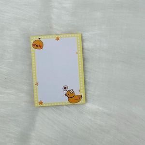 May include: A rectangular yellow sticky note pad with a white interior. The border is yellow with small white dots and orange stars. Cute cartoon duck illustrations are in the corners. The pad is on a white, fluffy surface.