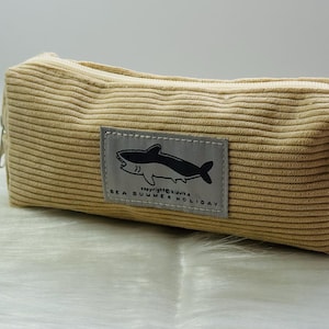 May include: A beige corduroy pencil case with a zip. A rectangular patch features a black and white shark illustration and the text "SEA SUMMER HOLIDAY". The case is rectangular and has a soft, textured appearance.