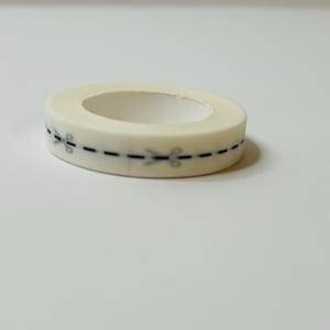 Peut inclure: Un rouleau de ruban adhésif washi blanc avec une ligne pointillée noire et de petites ciseaux imprimés dessus.
