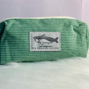 May include: A green corduroy pencil case with a white border and a shark graphic. The graphic says "copyright kisivita. sea summer holiday".