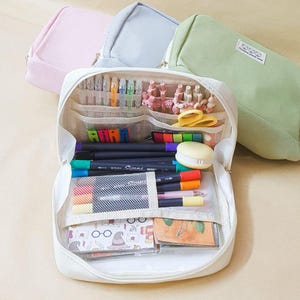 May include: A white canvas pencil case with multiple compartments and mesh pockets. The case is open and filled with various writing utensils, including pens, pencils, highlighters, and markers. There are also small decorative items, such as clips and a small cake.