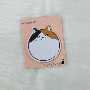 May include: A white, round sticky note with a cartoon cat design. The cat is orange, black, and white with a smiling face. The sticky note is on a pink and white grid background with the text "MENGOU" in black.