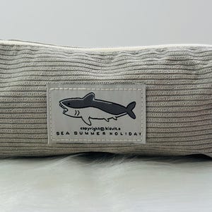 May include: A grey corduroy pencil case with a white zipper and a white patch featuring a black and white shark illustration and the text "SEA SUMMER HOLIDAY".