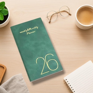 May include: A green weekly and monthly planner with gold lettering and the number 26, next to a cup of coffee, glasses, and a small plant. A notepad and flowers are also on the light wood surface.