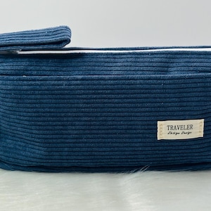 May include: A navy blue corduroy pencil case with a white zip and a small fabric label that reads "TRAVELER". The rectangular case has a handle and a pocket on the front.