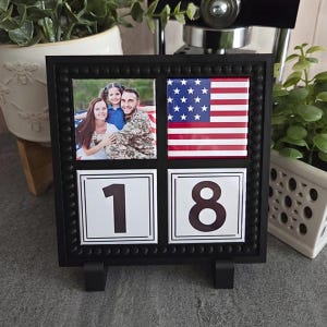May include: A square black frame with a beaded border containing four tiles. The top left tile shows a photograph of a family, the top right tile displays the American flag, and the bottom two tiles feature the large black numbers 1 and 8.