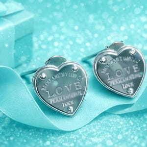 May include: A pair of silver heart-shaped stud earrings featuring the engraved text LOVE PERMANENT LOCK. Each earring has a polished metallic finish and a small circular accent at the top and bottom, presented alongside a light blue gift box and ribbon.