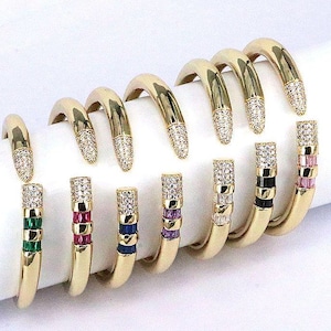 May include: Set of eight gold bangle bracelets with clear crystal accents and colorful stones. Each bracelet has a different color stone, including green, red, blue, purple, pink, black, and white.