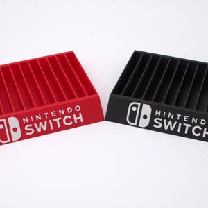 May include: Two rectangular game cartridge storage organizers, one red and one black, each featuring the white Nintendo Switch logo on the front face. Both organizers have multiple vertical slots for holding game cases in an upright position.
