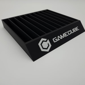 May include: Black GameCube game cartridge holder with multiple slots. The holder has a slanted front with the GameCube logo and text in white. The holder is made of a solid, matte black material.