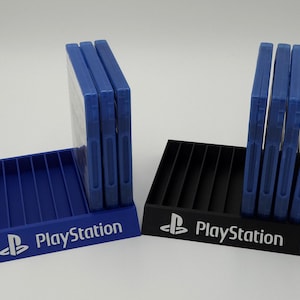 May include: Two PlayStation game organizers, one blue and one black, each holding several blue game cases. The PlayStation logo and name are printed in white on the front of each organizer. The organizers have a slotted design.