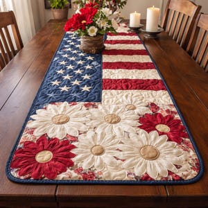 May include: A patriotic table runner featuring the American flag design with stars and stripes. The runner is adorned with large, fabric daisy flowers in red, cream, and tan. The edges are trimmed with a dark blue border. The runner is placed on a wooden table.