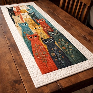 May include: A quilted table runner featuring a repeating pattern of stylized cats in various colors, including orange, yellow, and blue. Each cat has embroidered floral details. The runner is bordered with a white, quilted edge and is displayed on a wooden table.