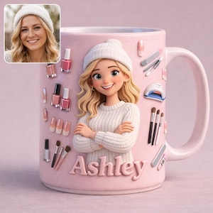 Könnte beinhalten: Rosa Keramiktasse mit einer Cartoon-Illustration einer Frau mit blonden Haaren und einer weißen Mütze. Die Tasse ist mit Nagellackflaschen, Nailart-Werkzeugen und dem Namen "Ashley" verziert. Ein kleines Foto einer Frau befindet sich in der oberen linken Ecke.