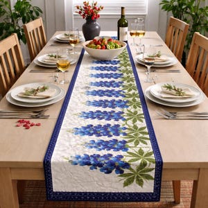 Könnte beinhalten: Ein Esstisch, der für eine Mahlzeit gedeckt ist, mit einem weißen Tischläufer mit blauen Blumenmustern und grünen Blättern. Der Tisch ist mit Tellern, Besteck und Weingläsern gedeckt. Eine Vase mit roten Blumen und eine Schüssel mit Obst sind ebenfalls vorhanden.