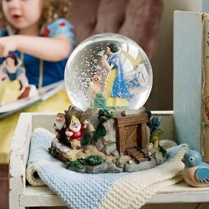 May include: A decorative snow globe depicting Snow White and the seven dwarfs. The globe is displayed on a shelf with vintage books, a blue bird ornament, and a small blanket. A child in a Snow White costume reads a book.