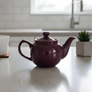 Puede incluir: Una tetera de cerámica color ciruela con tapa y asa a juego se encuentra sobre una encimera. Una taza blanca sobre un posavasos de madera y una pequeña suculenta en maceta también están sobre la encimera.