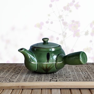 May include: A glossy green ceramic teapot with a side handle and a matching lid. The body of the teapot features a painted dark brown bamboo leaf design. The teapot has a short spout and a rounded shape, showcasing a smooth, reflective finish.