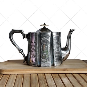 May include: A silver teapot with decorative engraved panels and a curved handle. The teapot is placed on a wooden chopping board, with a bamboo surface in the foreground and a white diamond pattern in the background.