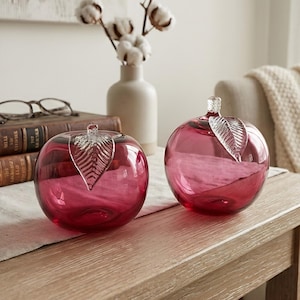 May include: Two glass apple-shaped vases in a deep pink colour, each with a clear glass leaf detail. The vases sit on a wooden table, with a neutral background featuring a vase of cotton stems and a cosy blanket.