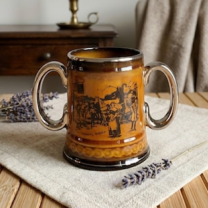 Könnte beinhalten: Eine braune Keramiktasse mit zwei silbernen Henkeln und einer detaillierten Schwarz-Weiß-Illustration. Die Tasse hat einen goldenen Rand und einen dekorativen Boden. Sie steht auf einer Leinen-Serviette auf einem Holztisch.