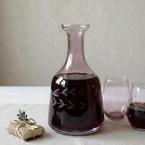 May include: A purple glass wine decanter and two matching stemless glasses, all filled with red wine. The decanter and glasses have a leaf design etched into the glass. A small wrapped gift with greenery is in the foreground.