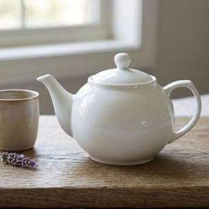 Könnte beinhalten: Eine weiße Keramik-Teekanne mit passendem Deckel und Griff steht neben einer beigefarbenen Tasse auf einem Holztablett. Ein Zweig Lavendel sorgt für einen Farbtupfer. Die Teekanne hat eine runde Form.