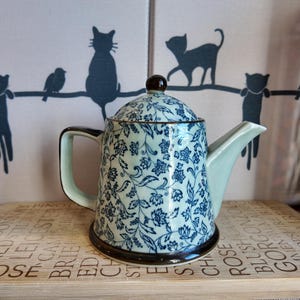Peut inclure: Une théière en céramique avec un couvercle, ornée d'un motif floral bleu sur un fond vert clair. La théière a une base, une poignée et un bouton de couvercle marron foncé. Elle repose sur une planche en bois avec du texte.