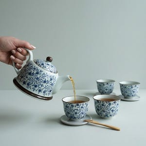 Könnte beinhalten: Eine weiß-blaue Teekanne mit floralem Muster gießt Tee in eine passende Teetasse. Drei weitere Teetassen und Untertassen sind sichtbar. Eine kleine Holzgabel liegt auf dem Tisch. Die Teekanne und die Tassen haben einen braunen Rand.