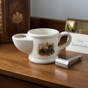 Puede incluir: Un tazón de afeitar de cerámica blanca con asa y compartimento lateral. El tazón presenta una ilustración de coche antiguo. Una maquinilla de afeitar de seguridad plateada descansa sobre una superficie de madera junto al tazón. El texto "The Essentials" está en una tarjeta.