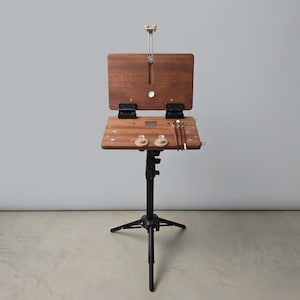 May include: A portable wooden pochade box easel with a dark brown finish, mounted on a black metal tripod stand. The easel features a hinged lid, a flat work surface with two clear circular knobs, and three paintbrushes resting on the right side.