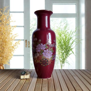 Puede incluir: Un jarrón de cerámica burdeos alto, con borde acampanado. El jarrón está adornado con un diseño floral con flores moradas y blancas. Los detalles dorados resaltan el diseño. Un artículo decorativo.