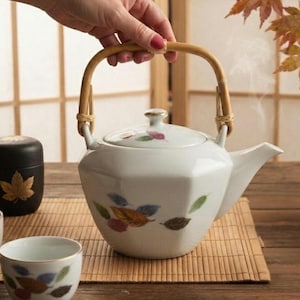 May include: A white ceramic teapot with a hexagonal body and a curved bamboo handle. The teapot features a painted pattern of colourful autumn leaves in shades of brown, blue, and green. It includes a matching lid with a small knob handle.