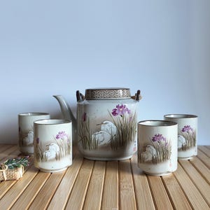 May include: A ceramic tea set featuring a teapot and four cups. The set is decorated with a hand-painted design of white birds, purple flowers, and tall grass. The teapot has a patterned lid and wooden handles. The cups are cylindrical.